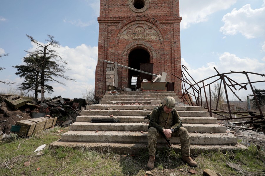 A soldier sits on the steps of a ruined structure, amid rubble.