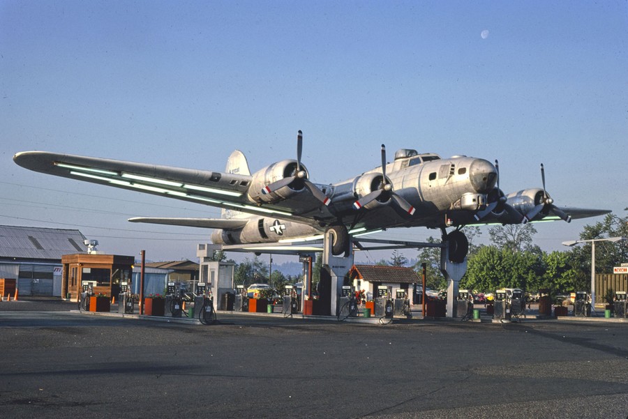 A large aircraft sits on struts above a gas-station filling area.