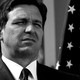 Black-and-white photo of Florida Governor Ron DeSantis squinting, standing in front of an American flag