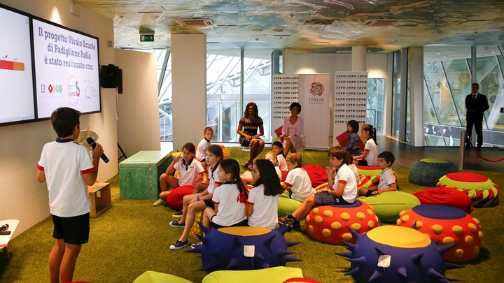 A group of elementary-school aged students sits on colorful chairs while looking at a powerpoint presentation. One boy holds a microphone and appears to be presenting.