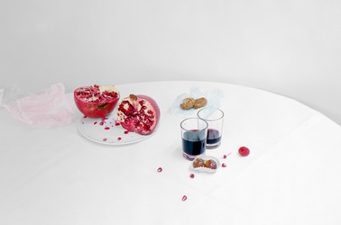 A white table with a pomegranate sliced down the middle on a white plate, and two glasses of pomegranate juice