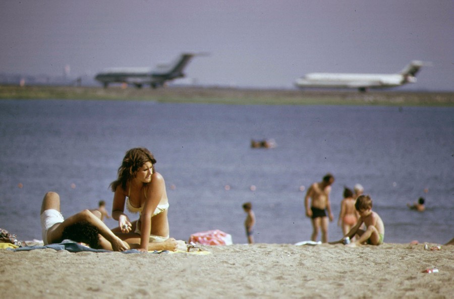 Several people relax on a beach with two airliners visible taxiing in the distance.