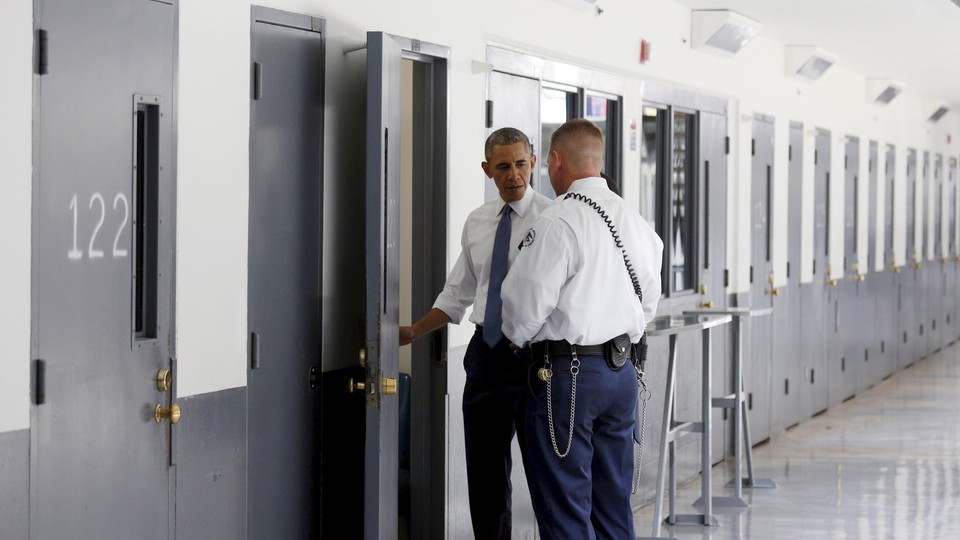 President Obama and a prison guard stand in front of an open door in a bright hallway, looking inside a prison cell.