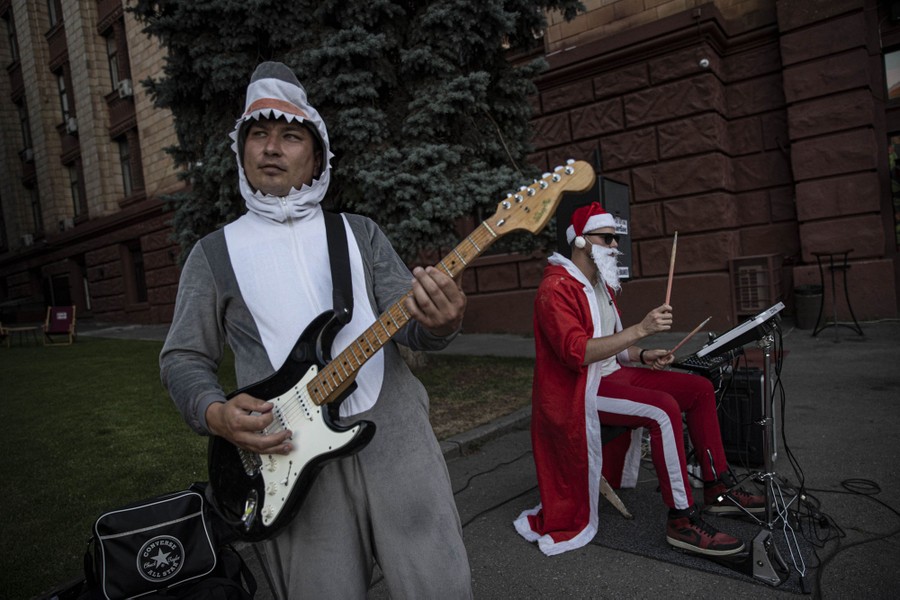 Musicians wearing costumes—one Santa Claus, the other a shark—perform on a street.
