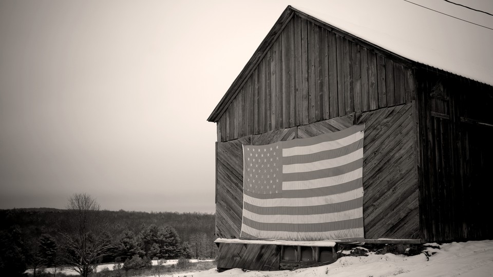 A black and white photo of a barn in the snow with a large American Flag hanging on it