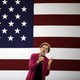 Elizabeth Warren, wearing a pink cardigan, speaks into a microphone onstage at an event. A giant American flag is displayed behind her.