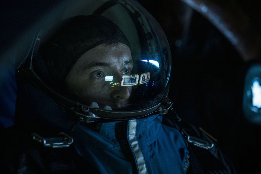A person wearing a space suit and helmet, seen in a darkened space, with several screens reflected in their visor.