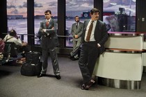 Photo of some suited men standing around an airport waiting area looking bored