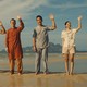Five people stand waving on a beach