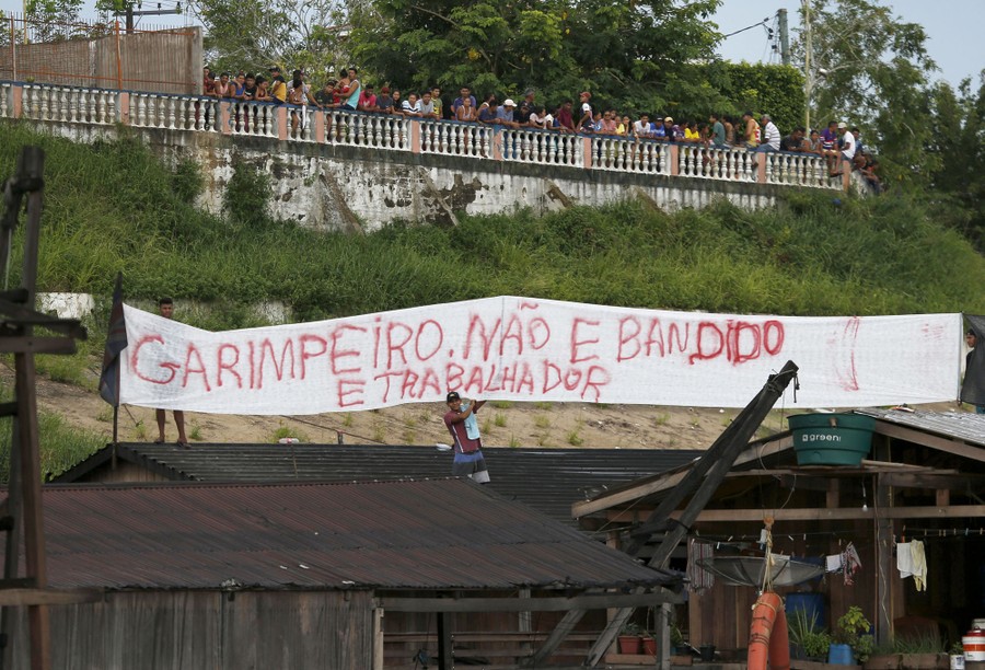 People gather on a terrace above barges docked on a river shore, with a banner below them that reads "Gold miners are not bandits; they are workers" in Portuguese.