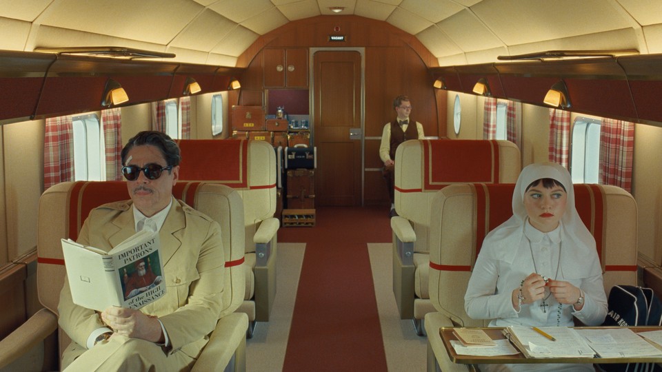 A color still from "The Phoenician Scheme" showing Benicio del Toro and Mia Threapleton in an airplane