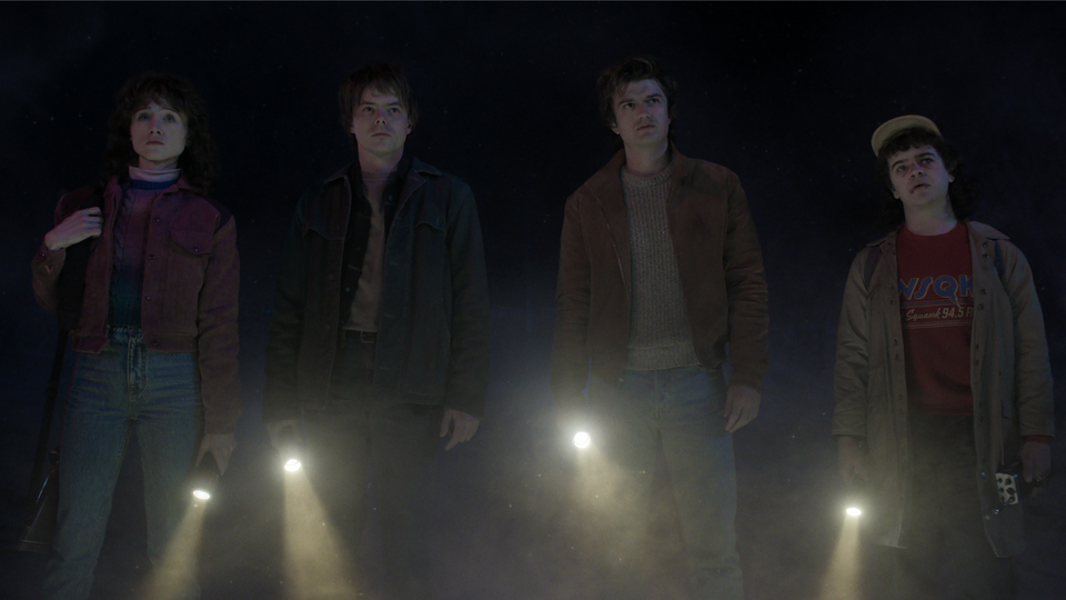 Nancy, Jonathan, Steve and Dustin from Stranger Things stand in a line, holding flashlights in darkness