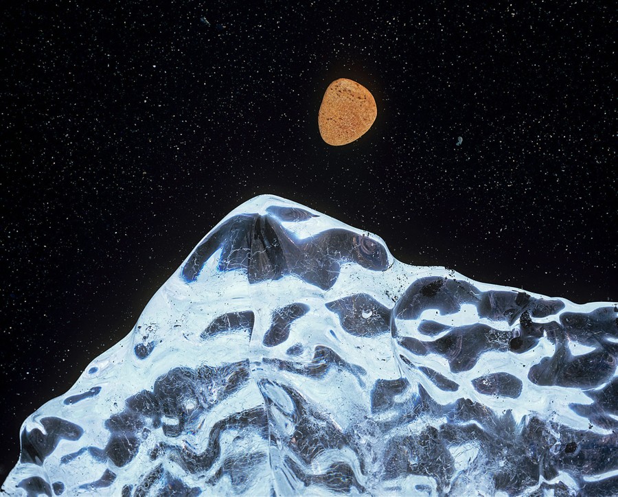 Looking down on an iceberg on a black sand beach beside a rounded orange pebble, creating a scene that looks like the moon over a mountaintop at night.