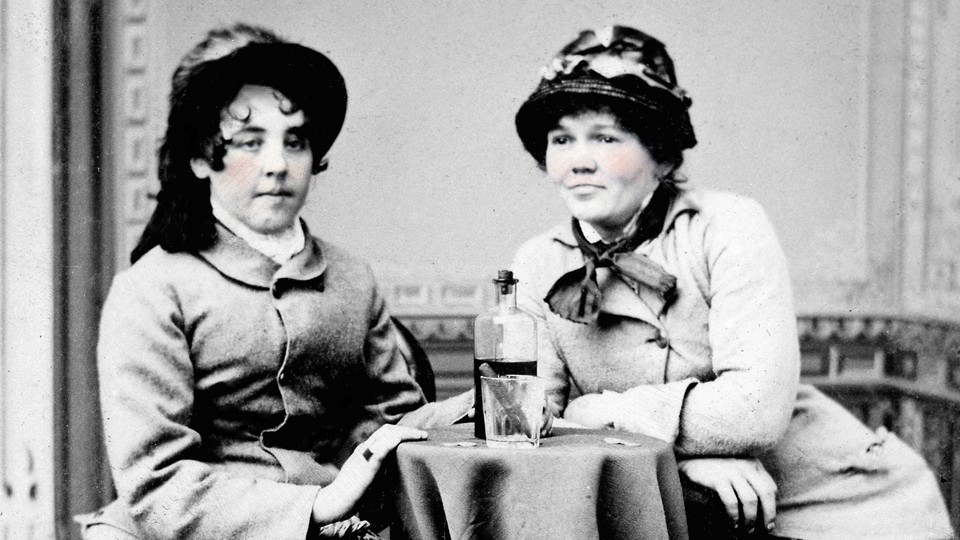 A black-and-white photo of two 19th-century women sitting at a table with a bottle of whiskey and a cigar