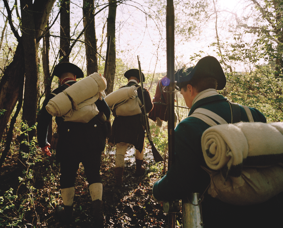 photo of reenactors as American soldiers wearing packs with bed rolls and hiking in a line through the woods towards Fort Ticonderoga