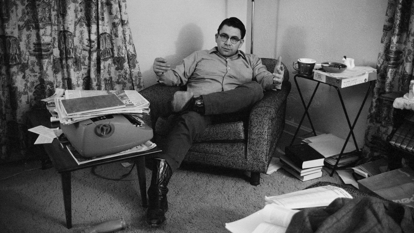 black-and-white photo of man wearing glasses sitting in living-room armchair by small table with typewriter and stacks of paper