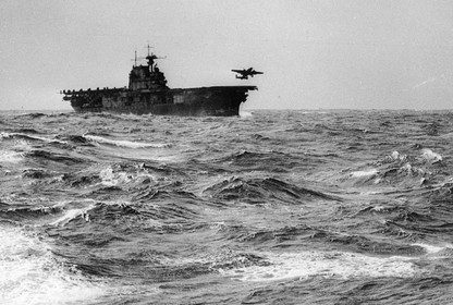 A bomber aircraft takes off from the flight deck of a US military ship in a turbulent ocean