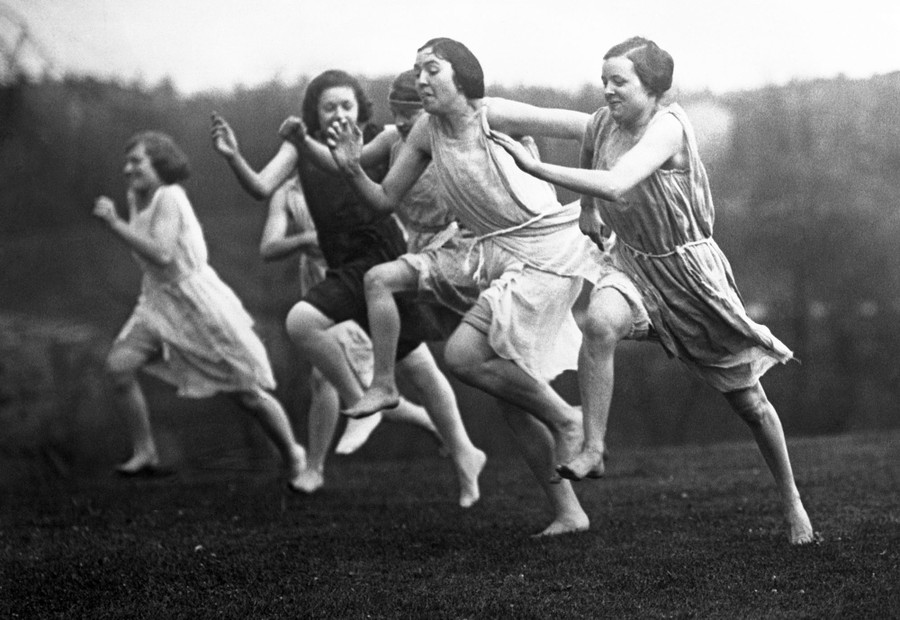 Young women race barefoot on a lawn, wearing loose garments meant to be those of ancient Greeks.