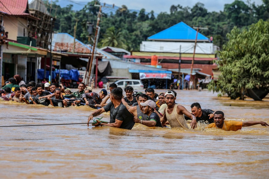 A line of people hold on to a rope as they wade through chest-deep flowing floodwater.