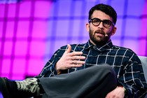 Photograph of Hasan Piker speaking with a microphone attached to his face. He is seated in a gray chair with his legs crossed.