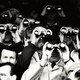 A black-and-white photograph of several men holding binoculars up to their faces to look at something far away