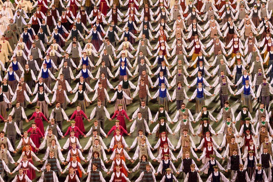 Hundreds of people in traditional clothing stand in rows, holding hands, during a performance.