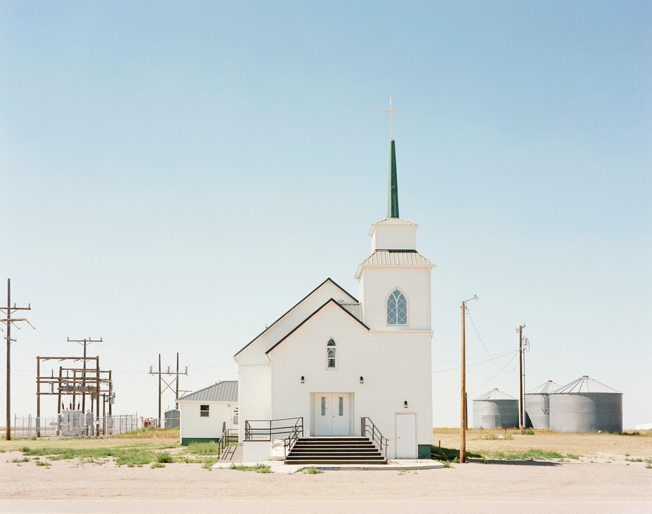 Photo couleur d'une église blanche avec un clocher, des lignes électriques le long du côté gauche et des silos de stockage sur la droite