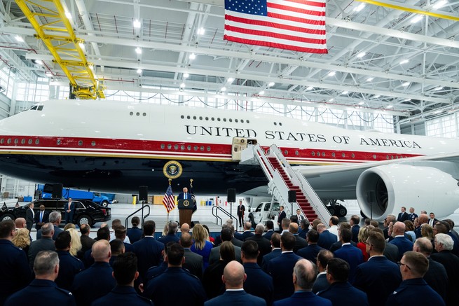 Photograph of Donald Trump giving a speech at a podium in an airplane hangar. An airplane with “United States of America” written on it is behind him.