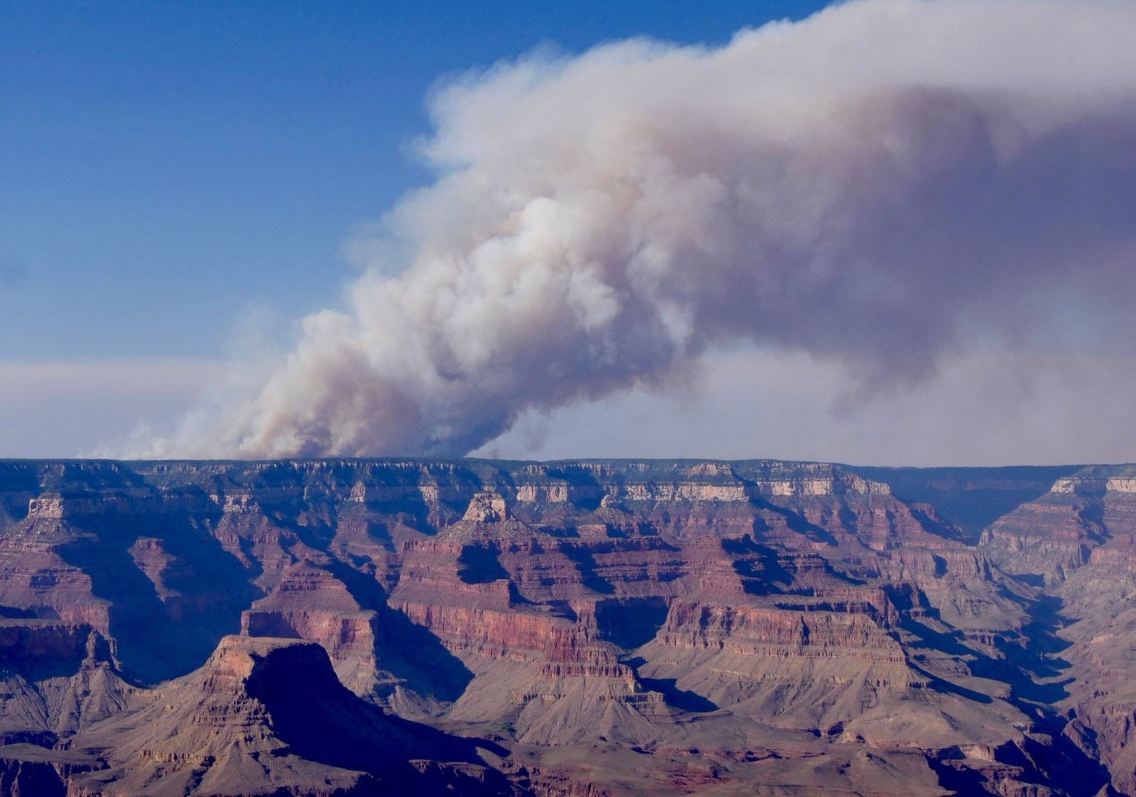 A distant plume of smoke rises above a wildfire on the far rim of the Grand Canyon.