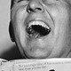 A black-and-white photo of a man throwing his head back in laughter while holding a book