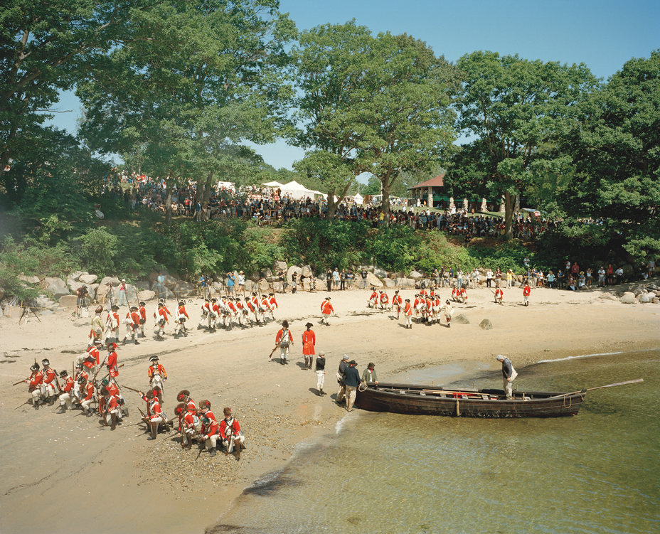 photo of a reenactment of the British arrival by sea, with wooden boat in shallow water being pulled onto beach and several groups of armed British troops on the sand, one kneeling with muskets, while a large crowd gathered under trees looks on from the green hill above and tents and a pavilion in the background