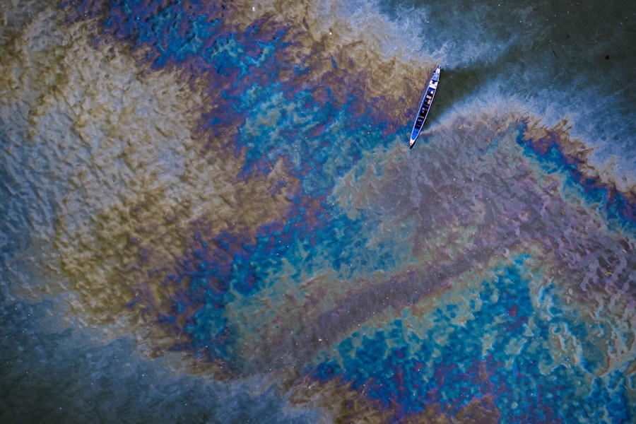 An aerial view of a small boat floating in a large rainbow-colored oil slick on the surface of a bay