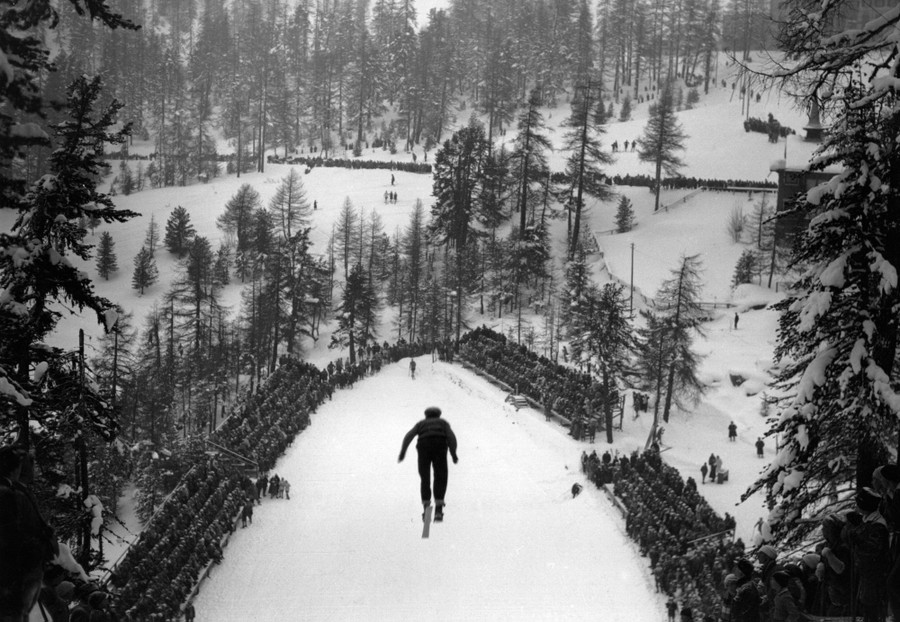 A rear view of a ski jumper in midair, with a crowd of observers below