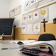 A teacher's desk is covered with papers in a classroom with a chalkboard and crucifix.