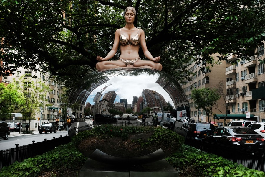 A realistic sculpture of a woman in a swimsuit sitting atop a large mirrored ball in a small green space in a road