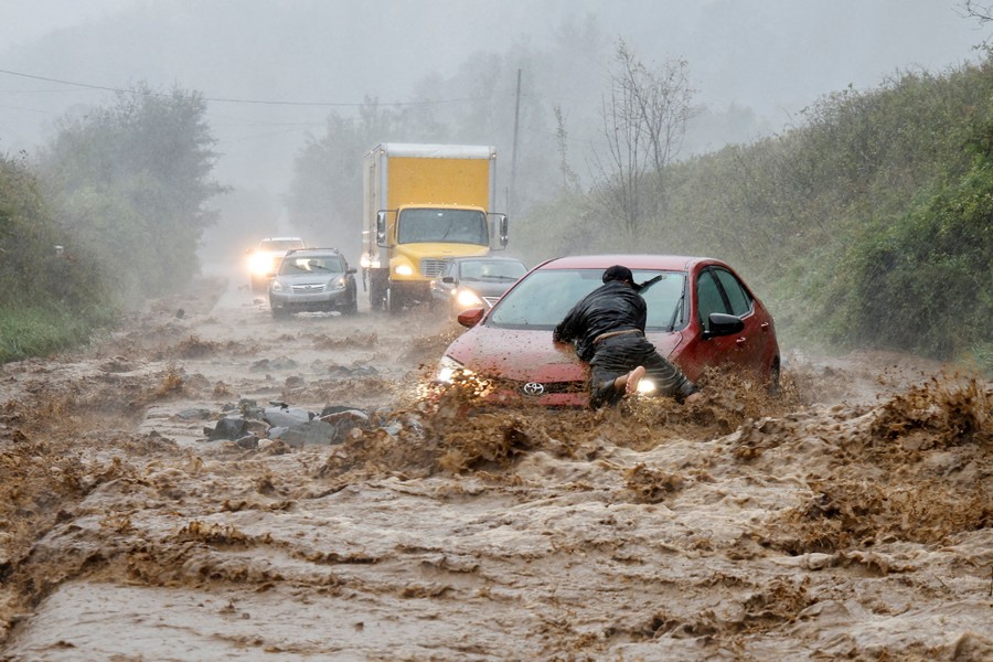 A person leans on the hood of a car that is surrounded by rushing floodwater on a road.