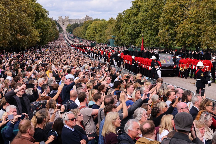 Thousands of people line both sides of a long road, watching a royal funeral procession pass by.