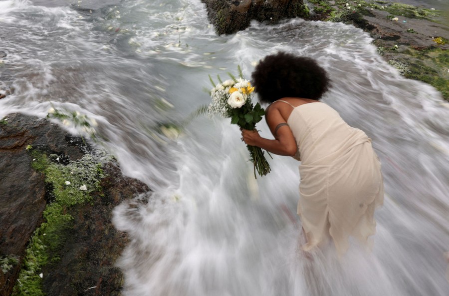 A person holding flowers leans down as an ocean wave rushes over their feet.