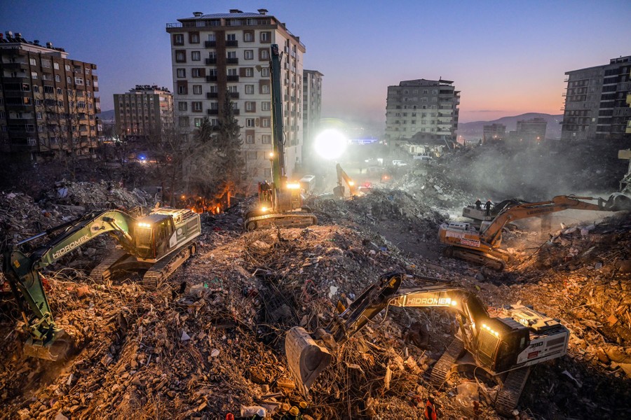 Several large excavators work on piles of rubble, amid residential buildings.