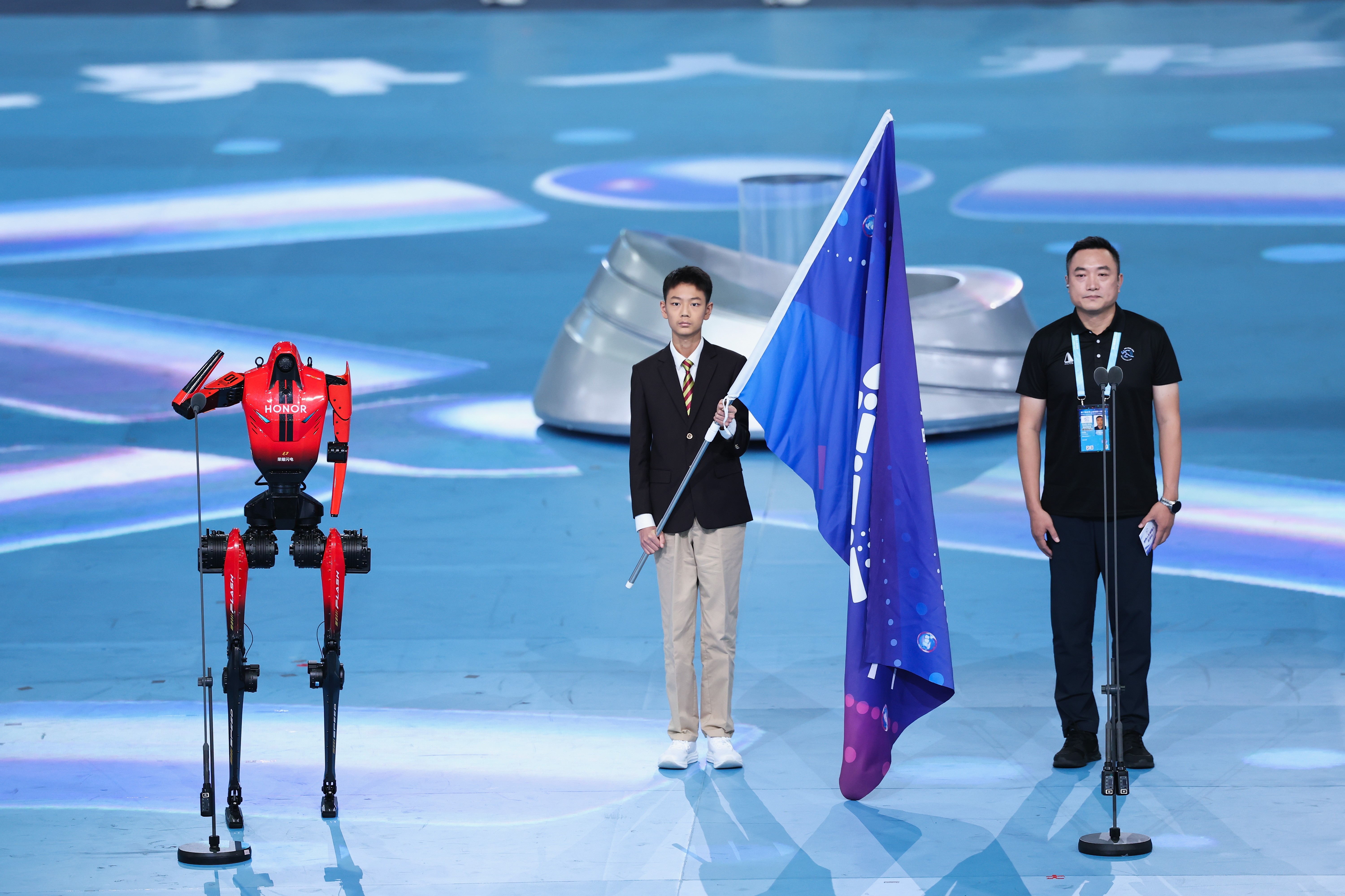 A humanoid robot stands at a microphone beside two actual humans, one holding a blue flag.