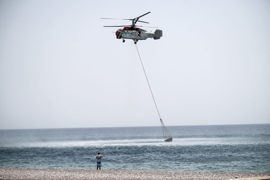 A helicopter flies close to the surface of the ocean, with a long cable hanging beneath it attached to a bucket that is being dipped into the water.