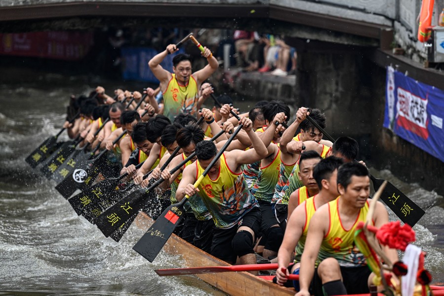 A couple of dozen people row vigorously, propelling a long boat forward in a canal during a race.