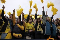 University of Michigan students hold yellow pom-poms and cheer in the stands of a football game. 