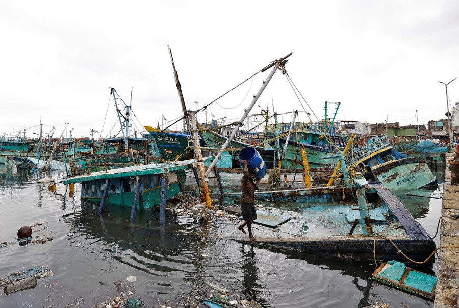A person carries a barrel away from a half-submerged boat in a harbor full of storm-damaged boats.