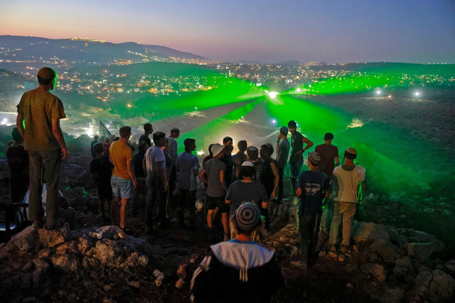 Settlers stand on a rocky hill at night, as people in the valley below shine green lasers toward them.