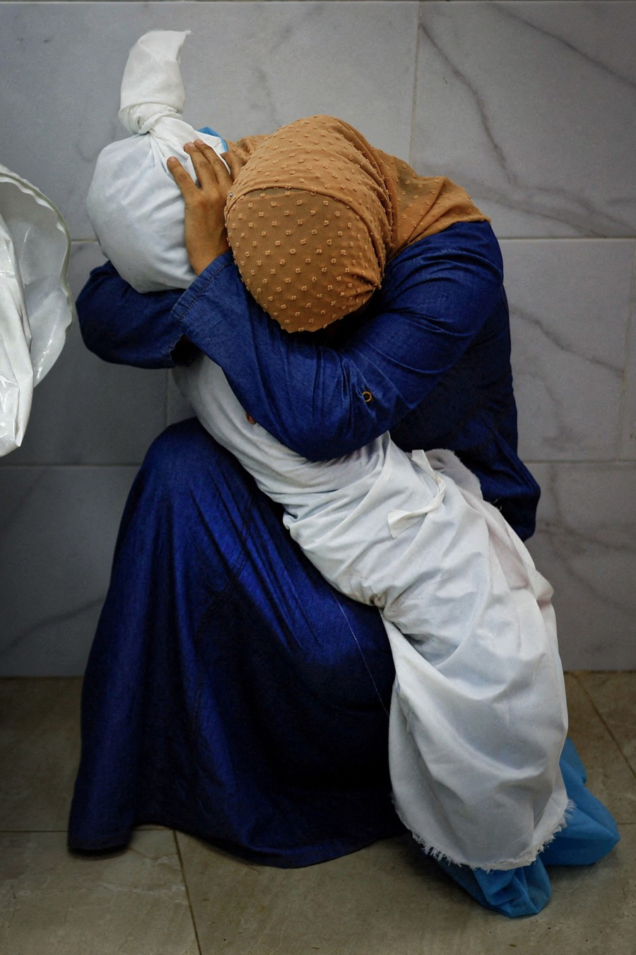 A woman kneels and puts her head down while embracing the body of a child, wrapped in white cloth.