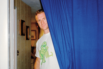 photo of man wearing white t-shirt with turtle logo and shorts looks out from behind a blue curtain inside a home