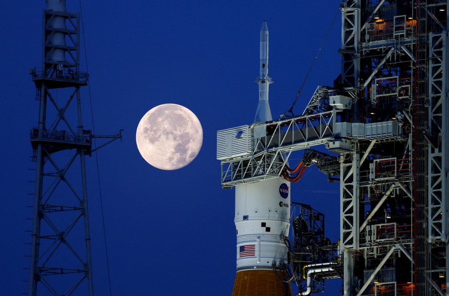The moon beyond the top of a large rocket that stands on a launch pad.