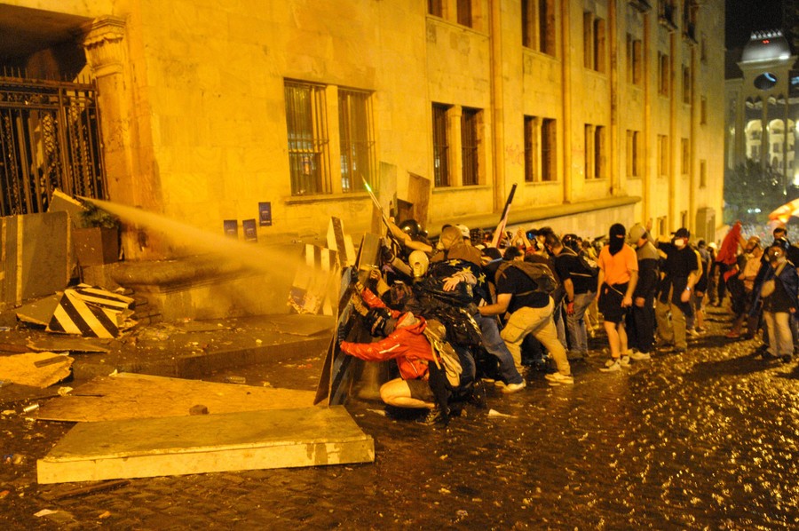 A crowd of protesters in a street hold up boards as shields against a water cannon directed at them from behind a gate.