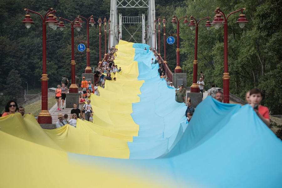 Dozens of people hold up an extremely long blue-and-yellow sheet of cloth, stretching down a pathway and over a bridge.
